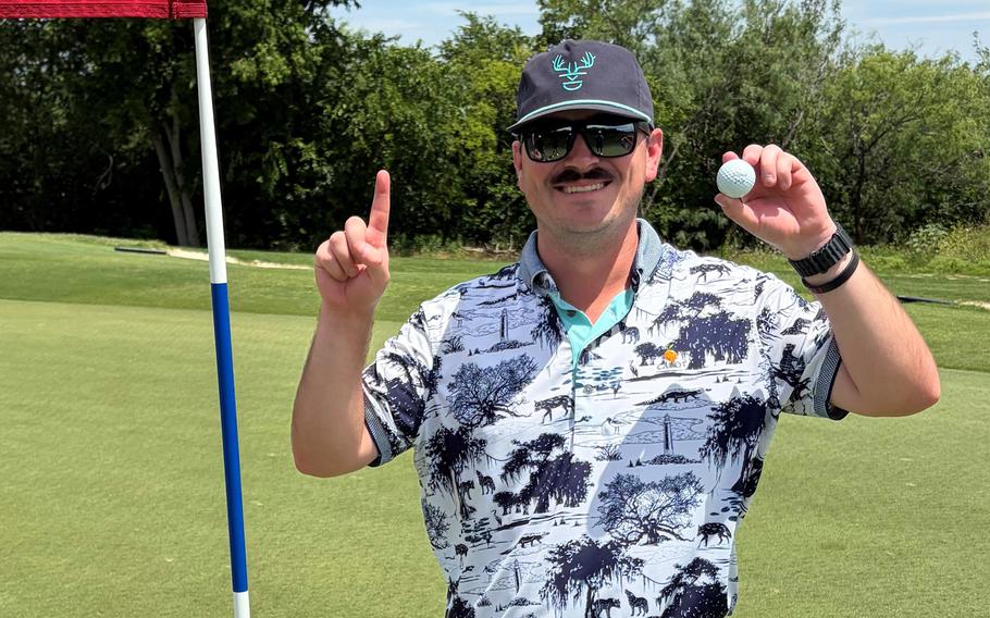 A man in a black and white flowered shirt, black hat and sunglasses poses on the green of a golf hole, with a ball in one hand and the other making a No. 1 gesture.