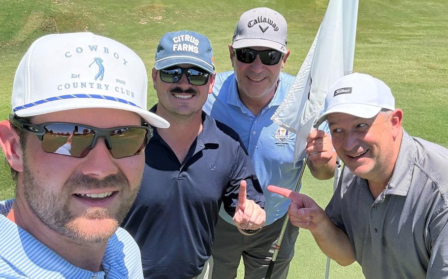 Four men in golf attire, hats and sunglasses pose on a golf green, with one holding a flag stick.