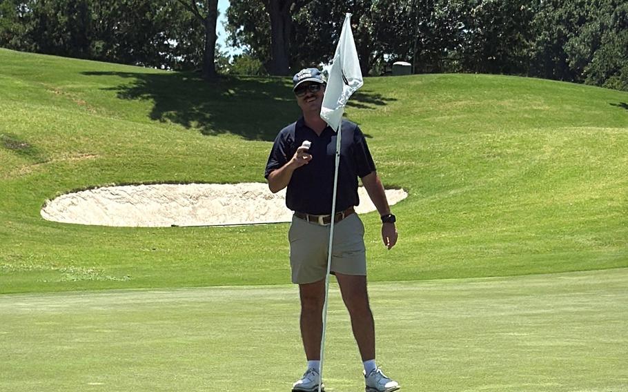 A man in a black golf shirt, khaki shorts, black hat and sunglasses poses on a golf green, standing behind the flag stick and holding up a golf ball.