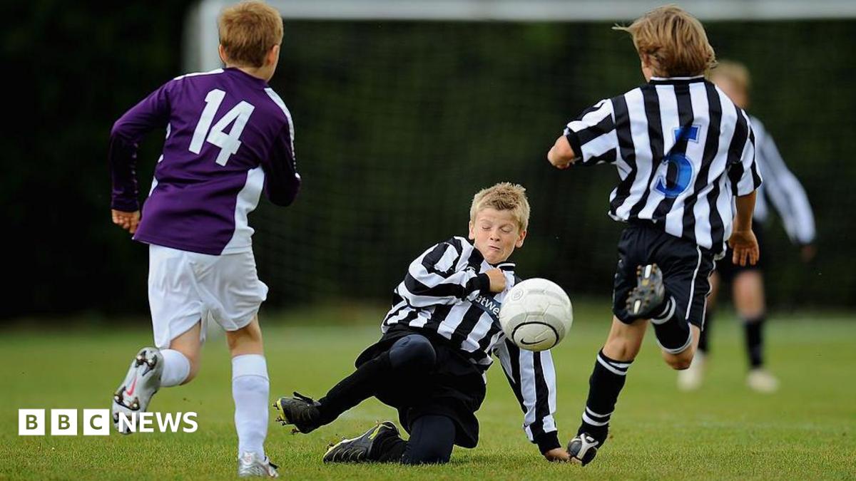 Three boys, playing football. One is on the ground, with a football near to his face, and his eyes closed. Two others have their backs to the camera and are running. Another child is blurred and in the distance.