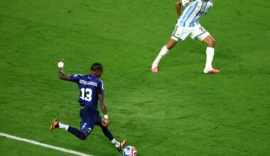 a soccer player in a navy jersey winds up to take a shot as a defender braces to block