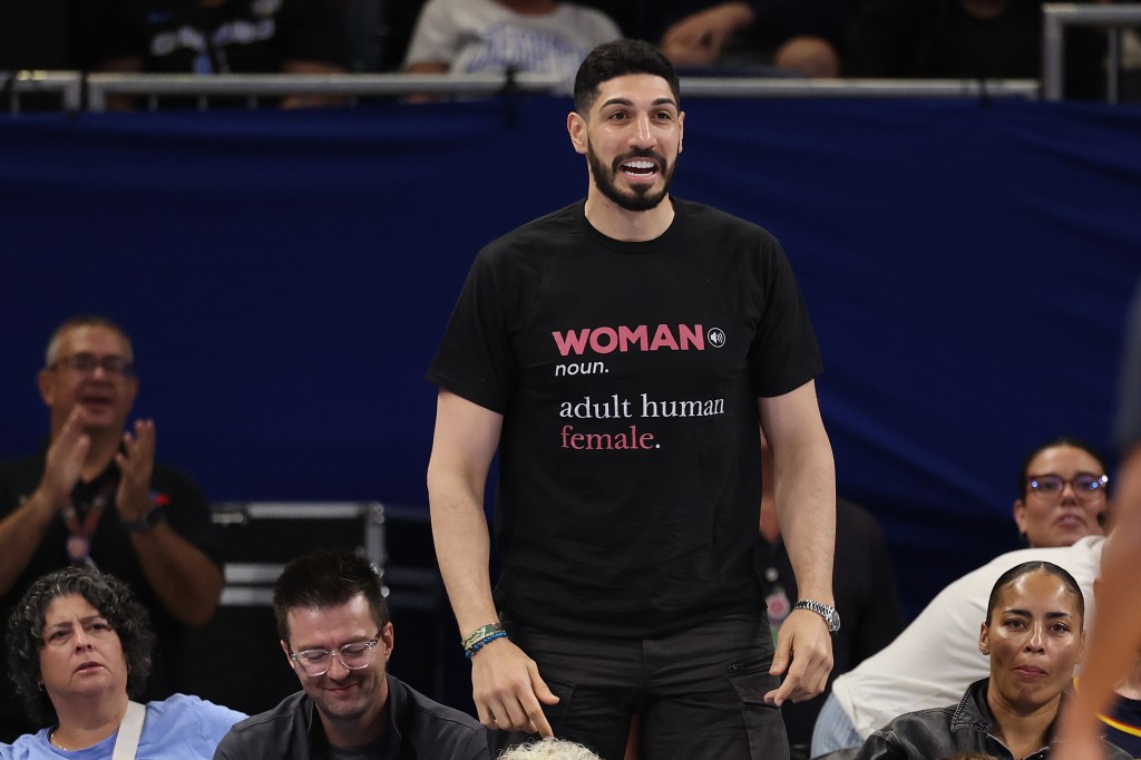 Enes Kanter Freedom, wearing a black shirt that reads "WOMAN noun. adult human female.", watches a game.