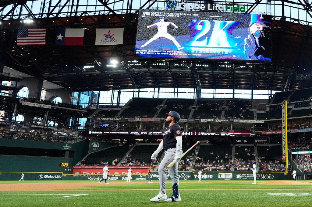 Baseball player Abimelec Ortiz walks to the dugout after striking out, with a jumbotron displaying "2,000 Strikeouts" for Jacob deGrom in the background.