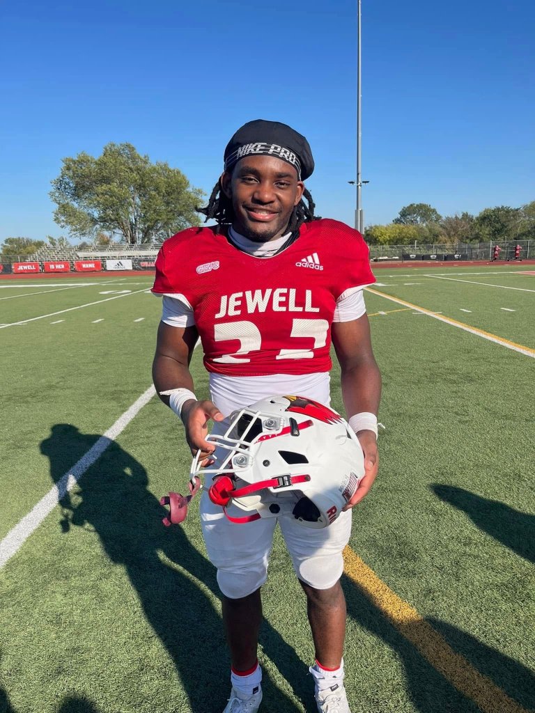 A football player in a red "Jewell 2" jersey holds a white helmet on a field.
