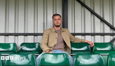 Dan sits on a green chair in a football stand, looking towards the camera. The seats are in a grey metallic stand and Dan wears blue jeans, a maroon top and brown jacket.