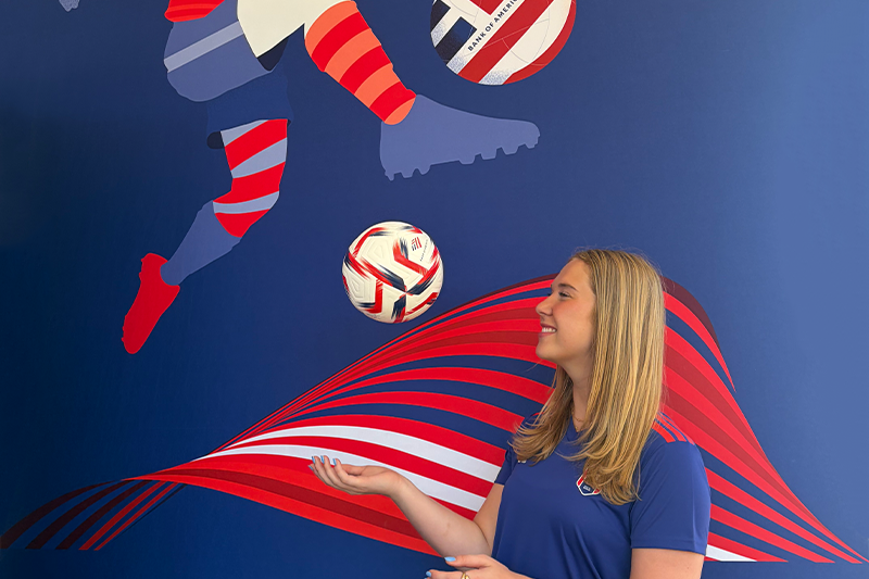 UNC alumna Emily Cancro ’26 smiles and poses beside a FIFA World Cup-themed mural and soccer ball while working as an event executive in Atlanta during the 2026 FIFA World Cup.
