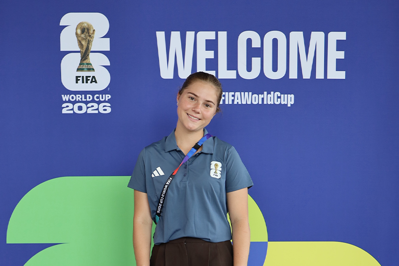 UNC junior Isla Mclaughlin stands in front of a FIFA World Cup 2026 welcome backdrop while working in transportation operations during the tournament in Atlanta.