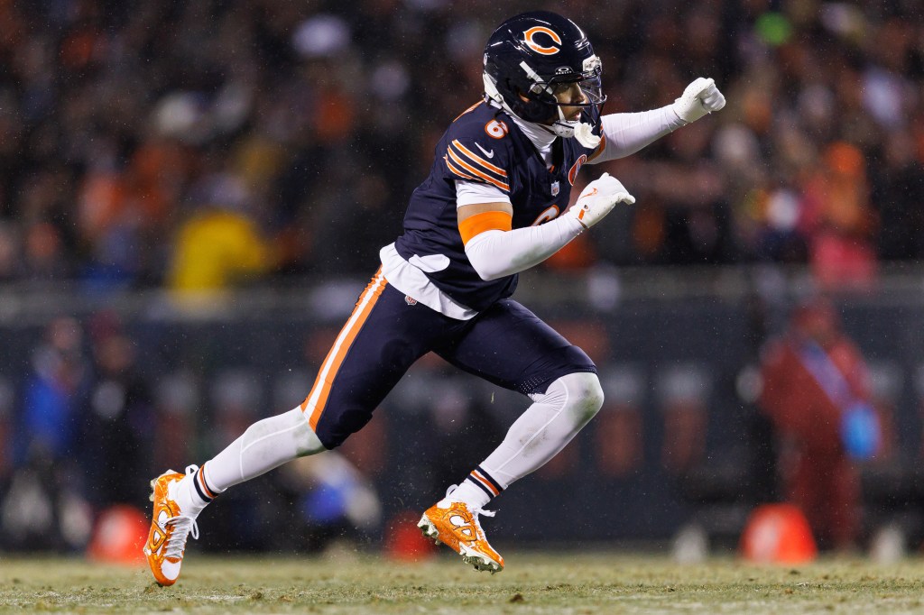 A Chicago Bears player in a dark blue uniform, white leggings, and orange and white cleats lunges forward on a football field.